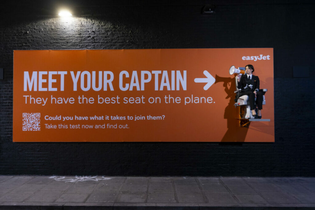 Text on a bright orange billboard, the same bold colour as easyJet's branding, reads "Meet your Captain. They have the best on the plane". It then points to Captain Sarah Ackerley who is dressed in her uniform holding a megaphone and is sat on an elevated pilot seat, which is part of the billboard. Underneath text next to a QR code reads "Could you have what it takes to join them? Take this test now and find out."