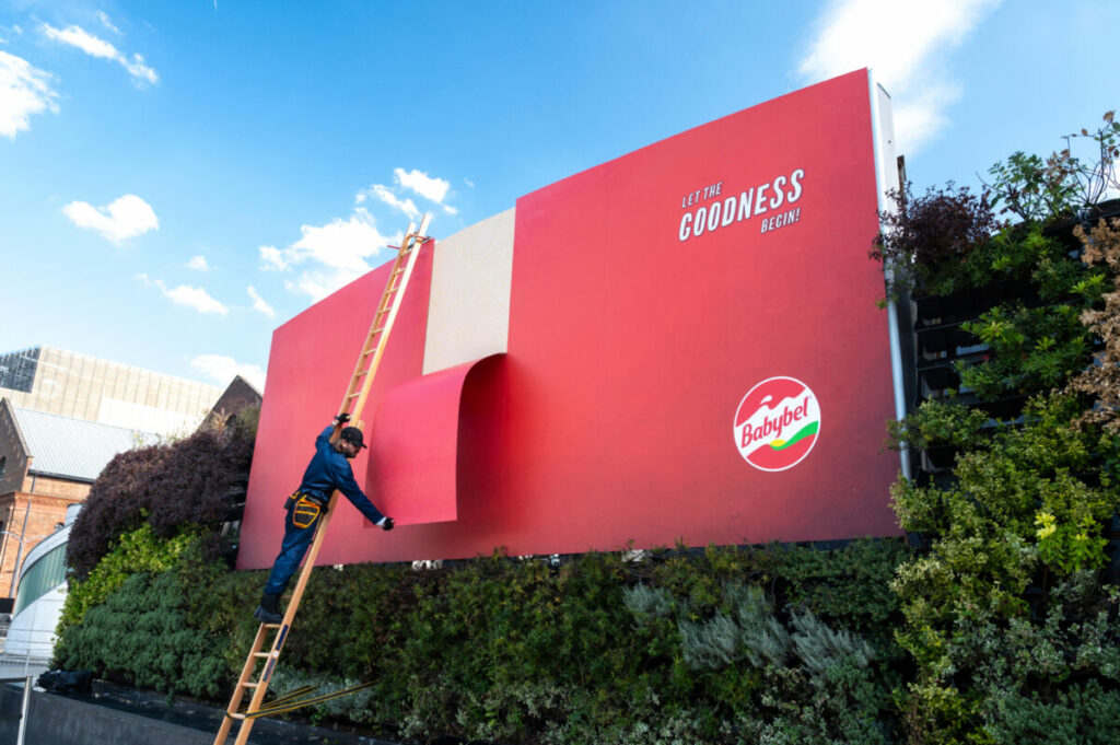 An actor on a ladder peels a giant red billboard (the Babybel branding) revealing white o represent cheese underneath under the tagline "Let the Goodness Begin" ahead of the Olympics. Babybel held its own iteration of an opening ceremony, as it unveiled giant peeling billboards ahead of the Olympics.