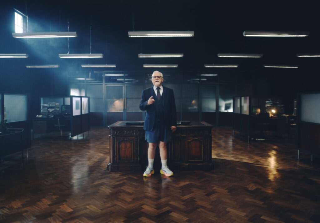 Brian Cox stands in a dark room in front of a desk in a shirt blazer tie running shorts and Asics running shoes.