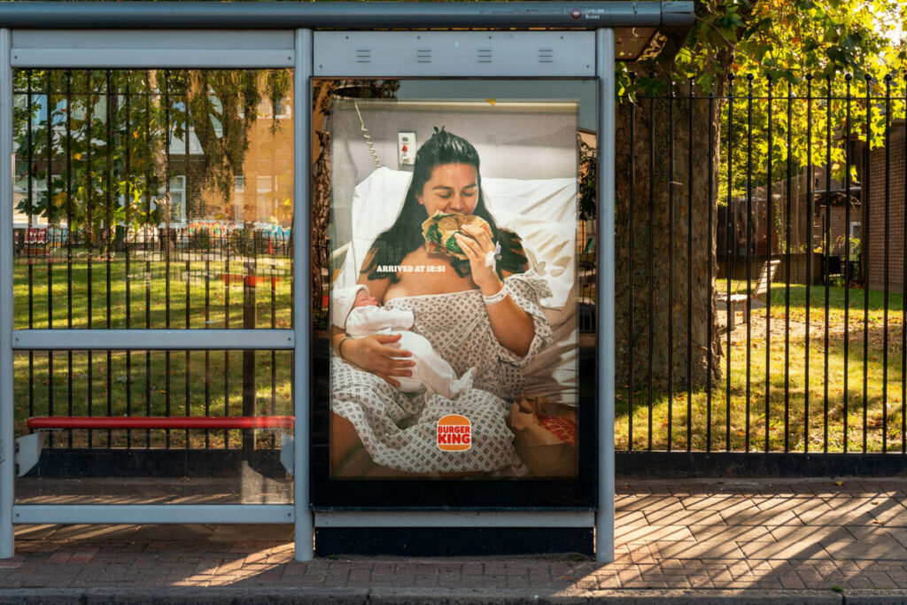 A new mum holding her sleeping new-born baby wearing a hospital gown munches ravenously on a Burger King burger.